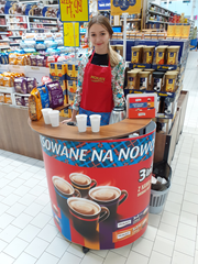 Uczennica na stoisku do promocji sprzedaży zachęca klientów do degustacji kawy w hipermarkecie podczas praktyki zawodowej.